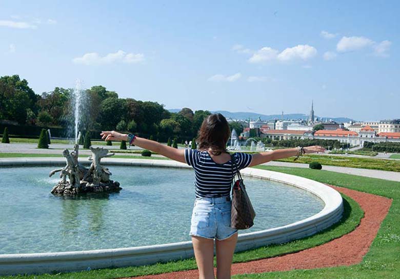 A youg girl standing in front of a fountain wirh a beautiful panorama of Vienna